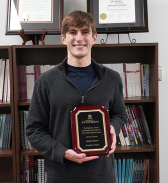A man holds a plaque in hand. It has a brown wooden frame surrounding a gold outline and black surface. It gold text it reads "The Dennis A. Pruitt Outstanding Advocate for First-Year Students Award 2026 Recipient Andrew Pana" with the USC logo overtop.