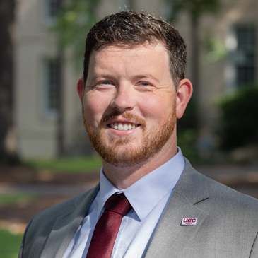 Scott McDonald stands on the USC Horseshoe wearing a gray jacket with a garnet USC pin