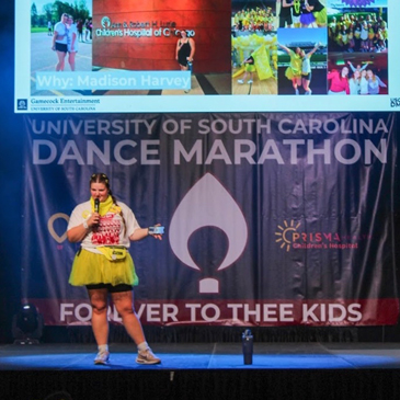 A student wearing a bright yellow tutu and a white Dance Marathon shirt stands on a stage speaking into a microphone. Behind her is a large banner reading “University of South Carolina Dance Marathon” with “For the Kids” and a Prisma Health logo, and a screen above displays photos and the heading “Why: Madison Harvey.”