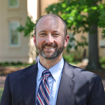 Portrait of Wright Culpepper wearing a navy suit, light blue shirt and striped tie, standing outdoors on the University of South Carolina campus.
