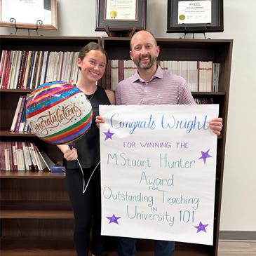 Wright Culpepper stands with peer leader Kate Heller holding a congratulatory balloon and a sign recognizing his M. Stuart Hunter Award for Outstanding Teaching in University 101.
