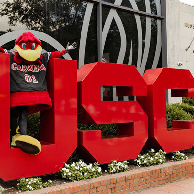 Gamecock mascot stands inside of a red-colored letter U with letters S and C to his left.