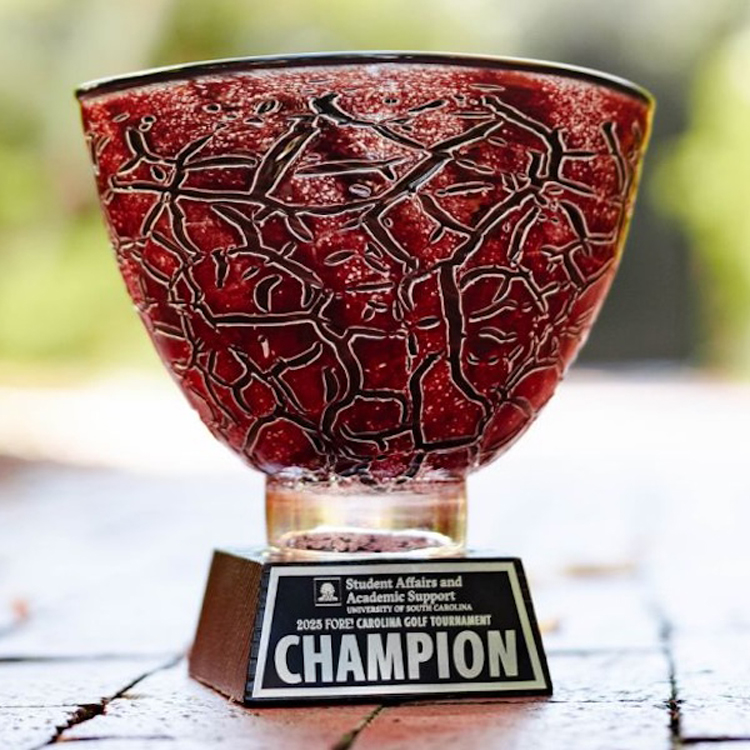 A close-up of a red, textured glass trophy bowl mounted on a wooden base with a plaque reading “Student Affairs and Academic Support, University of South Carolina — 2025 Fore Carolina Golf Tournament Champion,” photographed outdoors on a wooden surface with a softly blurred green background.