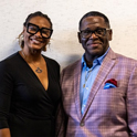 Woman and man, both wearing dark-framed glasses, stand in front of a beige wall.