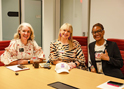 Jennifer Jackson, Jennifer Cristina and Shonta Sellers sit side-by-side in garnet chairs around a wooden table. A white and purple food fight hat sits atop the table.
