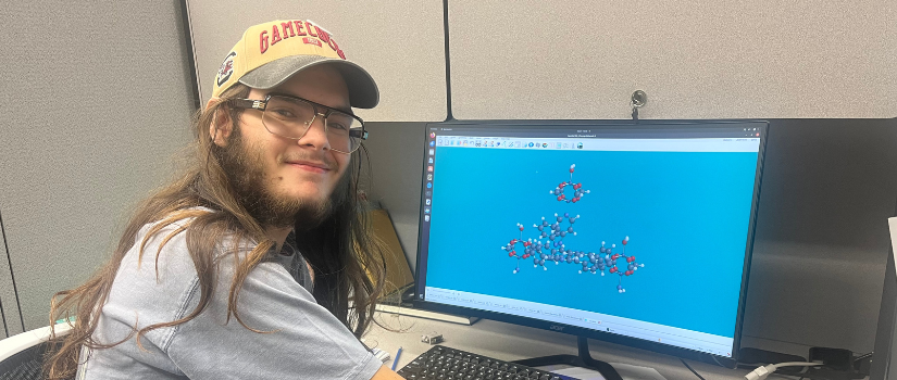 A person with long hair and glasses sits at a cubicle desk, smiling at the camera while using a computer that displays a 3D molecular structure on the screen.