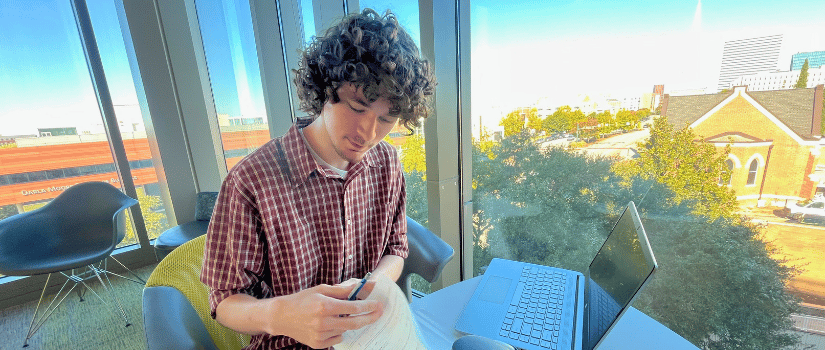 A person with curly hair sits at a table by a large window, reviewing printed documents with a pen while a laptop and water bottle rest on the table, with a campus view visible outside.