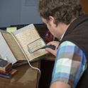 Over the shoulder view of a person using a handheld magnifying glass to read handwritten text in a manuscript. The book is resting in a book cradle on the table.