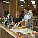 A librarian assists a patron at the Thomas Cooper Library reference desk. Both people are standing at the counter, looking at computer monitors.