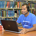 Person sitting at a library table wearing headphones and looking at a laptop. Library shelves filled with books in the background. 
