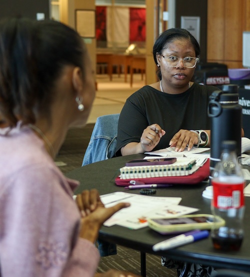 two people sitting at table with notebooks and items, speaking together