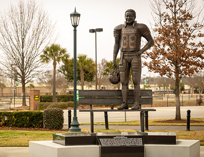 Statue of George Rogers in his football uniform with then number 38 on the jersey, standing on a bench outside of Williams-Brice Stadium.