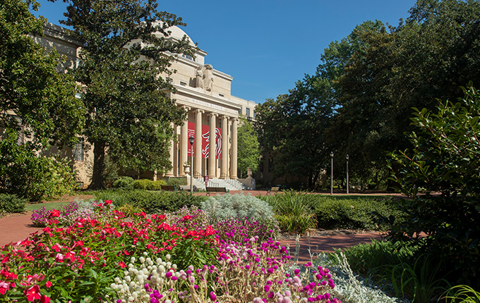 McKissick Museum sits surrounded by green trees and colorful flowers with garnet banners between it's stone columns. 