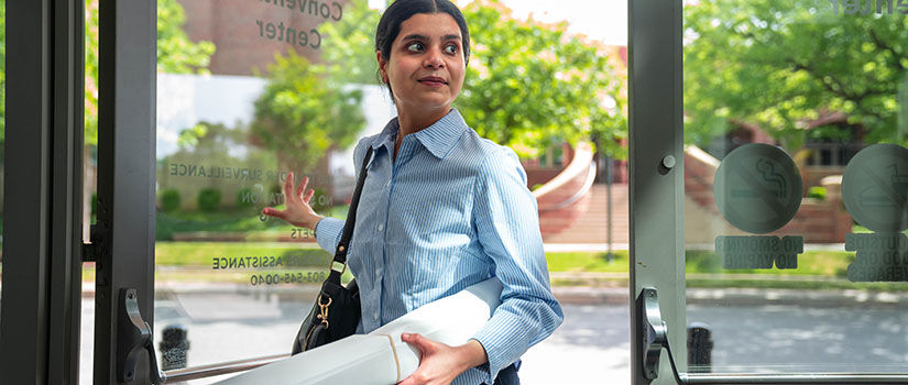 A woman wearing a blue striped button-down shirt enters a glass doorway at the Columbia Metropolitan Convention Center while holding a rolled-up Discover USC poster and a reusable water bottle. She carries a shoulder bag and looks to her left as she steps inside, with greenery and a walkway visible outside behind her.