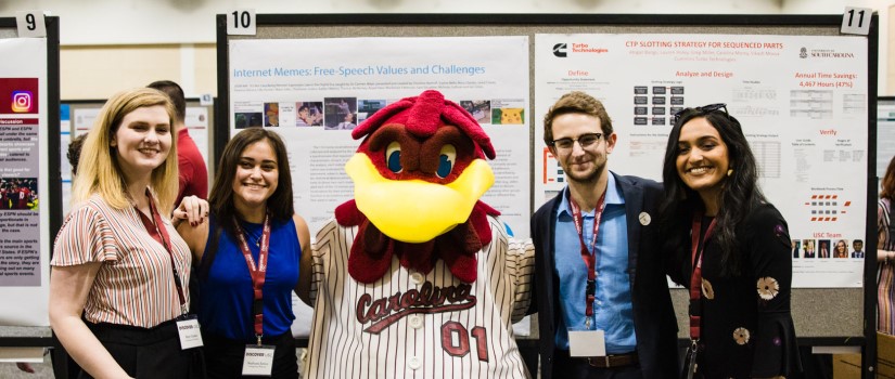 Photo of Cocky posing with presenters in front of a scholarly poster at Discover UofSC 2019.