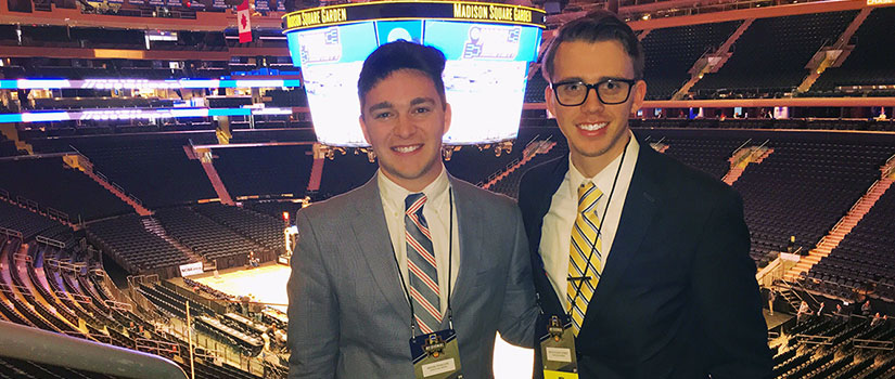 Two male students, wearing suits and ties and ID badges on lanyards, pose in Madison Square Garden arena, with the Jumbotron in the background
