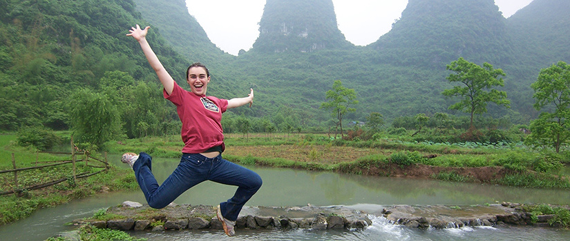 Female student on study abroad trip leaps into the air with arms extended, in front of mountains and a stream