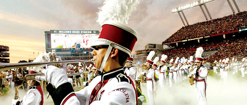 Carolina Band members in uniform and playing their instruments as they stand on the field at Williams-Brice Stadium, with the scoreboard and stands full of fans in the background