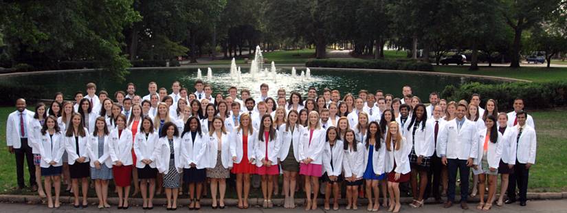 About 100 medical students, each wearing a white lab coat, stand in rows to pose as a group in front of an outdoor fountain