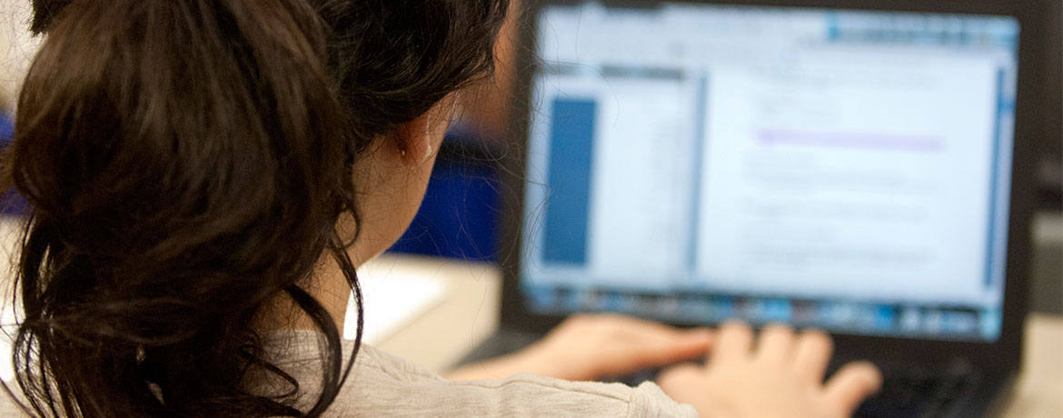 Close-up of the back of a female student's head and right shoulder, and her hands are on the keyboard of a laptop computer showing a document on its screen