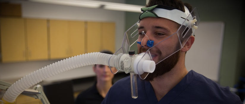 Male Public Health student in dark-colored scrubs demonstrates the use of a breathing apparatus that he wears on his head and face