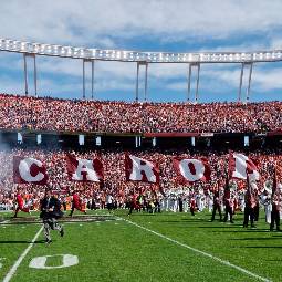 Football field on game day with cheerleaders carrying flags that spell out Carolina and fans filling the stands in the background