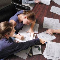 Two female students sitting at a work table covered in class notes, passing papers back and forth while they study