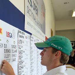Male student in green baseball cap working at a U.S. Open qualifier tournament, posting scores on a wall poster