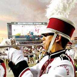 Close-up of Carolina Band member in uniform, playing trumpet, on football field with Williams-Brice Stadium scoreboard in background