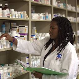 Pharmacy student in white lab coat checks shelves stacked with medicine bottles while holding open a notebook in her other hand