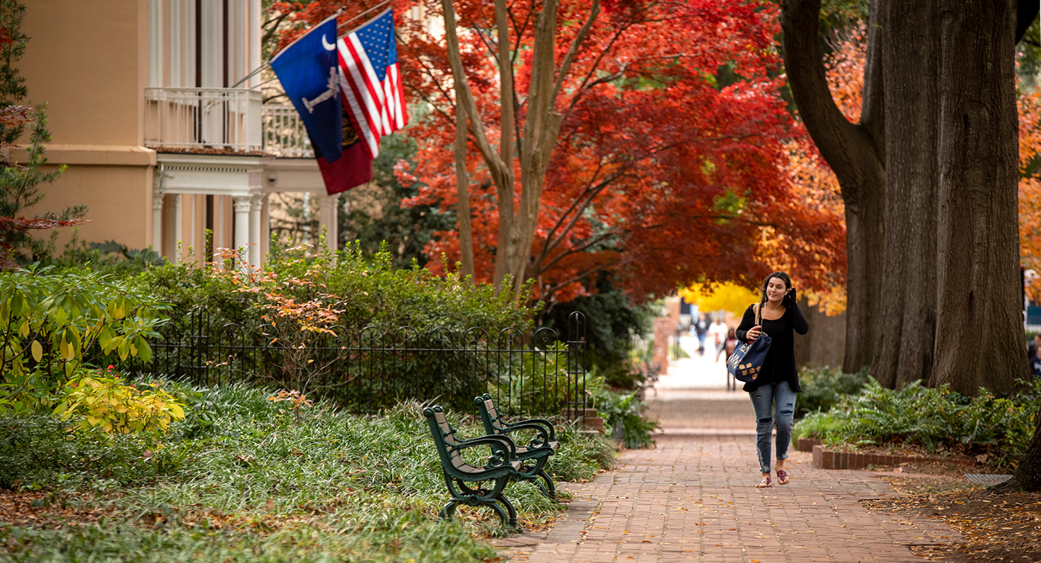 Student walking down a brick pathway with benches and the President's House with the State, American and university flag flying. 