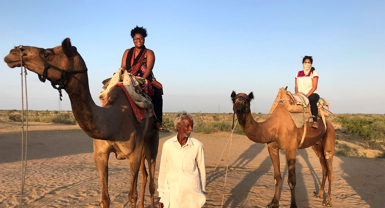 Andrea Bowman and another student sit on camels in the desert with guide leading them.