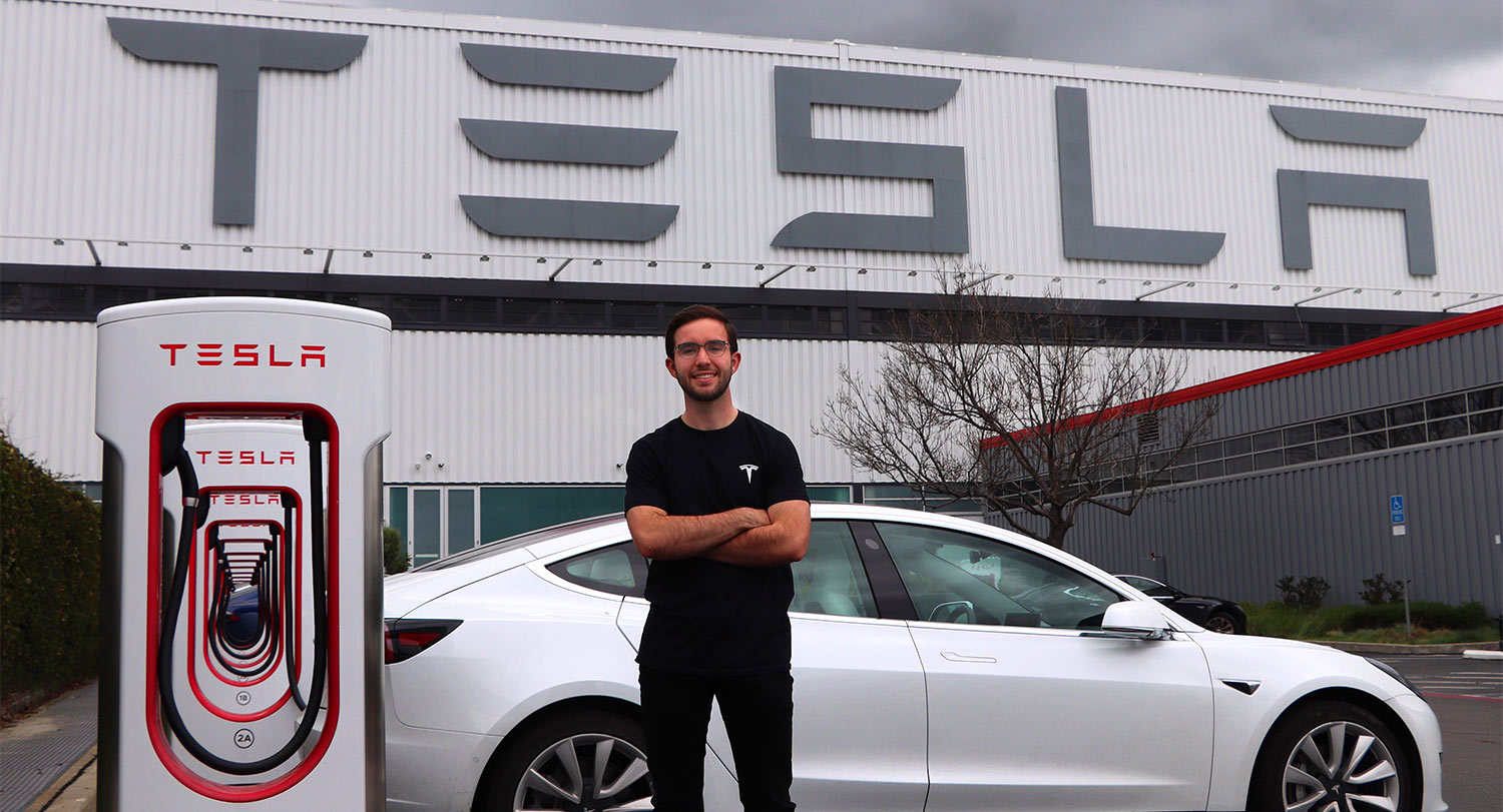 Brian Youngblood standing in front of an electric car with the Tesla logo on the building behind him and on the power stations beside the car. 