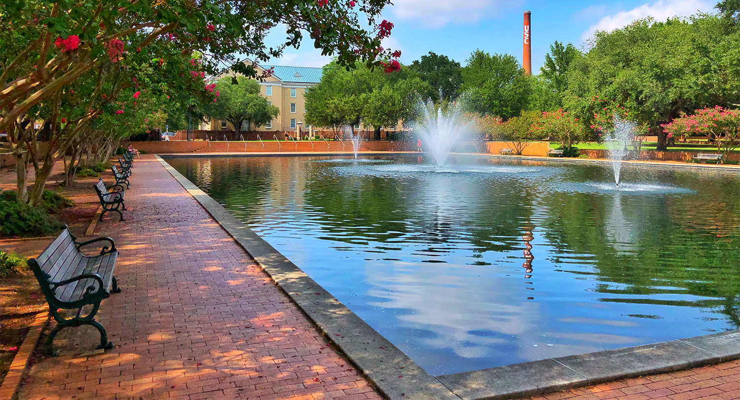 The reflecting pool with fountains and benches surrounding it and the USC smokestack in the background.
