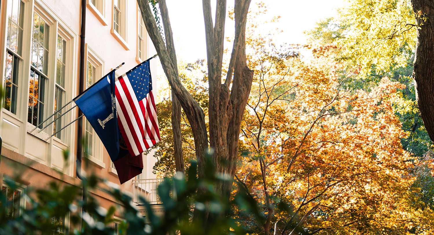 The American Flag and the South Carolina State Flag hang off the side of the President's House surrounded by fall trees on the Historic Horseshoe.