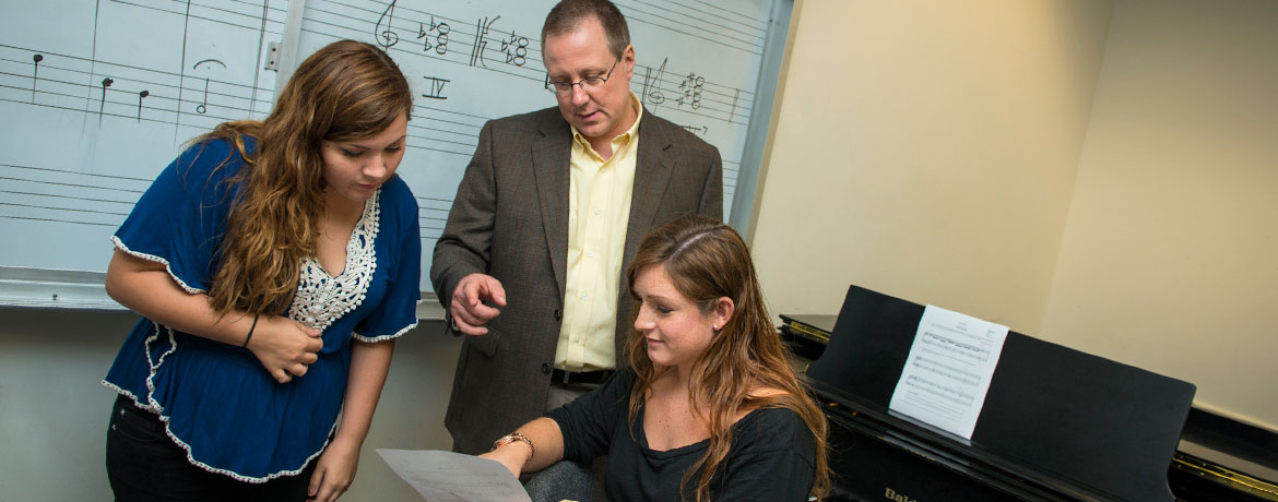 two music students consult with a professor in a practice room with piano and whiteboard