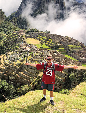 Student wearing a garnet football jersey standing with his arms stretched out in front of Machu Picchu.
