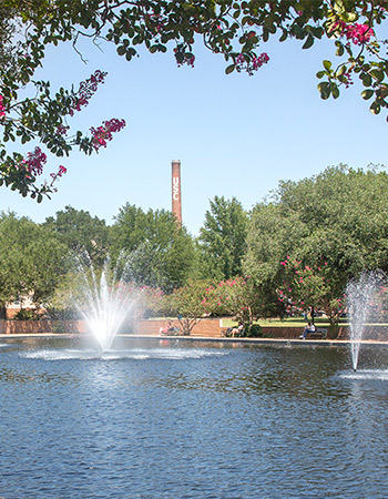 Running fountains in the reflecting pool surrounded by brick pathways and benches with a canopy of trees and the USC smokestack in the background.
