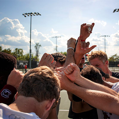 Students on a tennis court in a huddle with their arms raised together. 