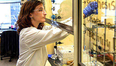 Student working in a chemical hood in a lab wearing a lab coat and safety equipment. 