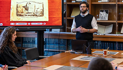 A professor standing in front of a large conference room table with a historical document on a screen beside him.