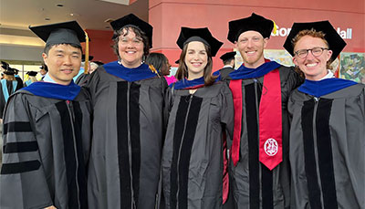 Group of five alumni in graduation gowns and doctoral hoods at a commencement ceremony.
