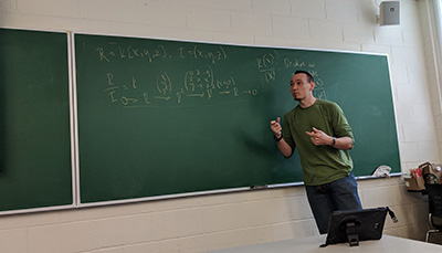 Student explaining something in front of a chalkboard with equations on it.
