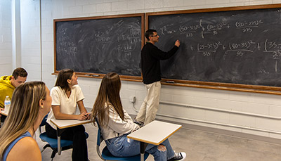 Students observing professor writing equations on blackboard in classroom.