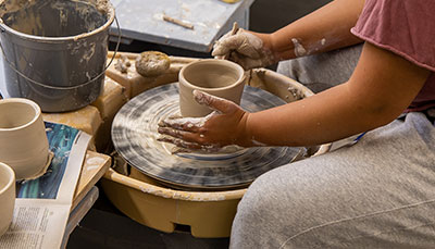 A student sitting at a pottery wheel forming a clay bowl. 