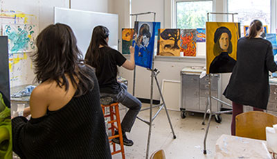 Students sitting in an art studio painting at on easels. 