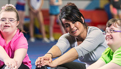 A female physical education teacher is seated with two female students and all three are stretching.