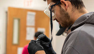Person wearing safety goggles and gloves holding a small piece of equipment in their hands.