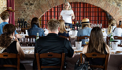 A person talking in front of a group of people sitting at tables at a wine tasting.
