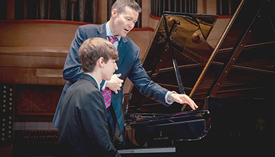 Professor giving instruction to a student sitting at a baby grand piano. 